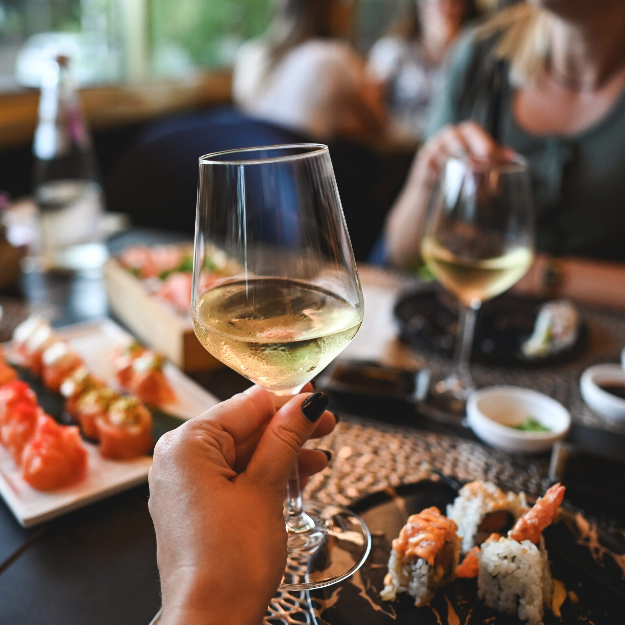 Hand holding white wine over sushi spread